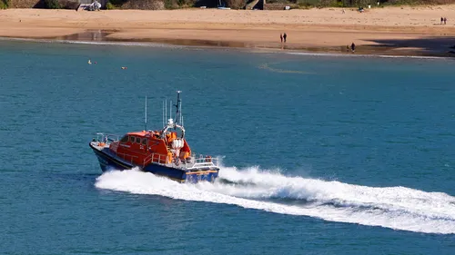 RNLI The Baltic Exchange speeding towards Salcombe Harbour - close This landscape photograph captures the RNLI lifeboat, The Baltic Exchange, speeding towards Salcombe Harbour in Devon, England, during the early afternoon in the spring. The vivid orange lifeboat is creating a distinct white wake as it moves quickly across the blue waters, illustrating its role as a vital transport vessel for emergency response. Visible in the background is the sandy shoreline of Salcombe, a well-known popular landmark in the United Kingdom. Small groups of people can be seen walking along the beach, which further emphasizes the everyday integration of RNLI lifeboats into coastal life in Devon. Boats such as The Baltic Exchange are essential for maritime safety and rescue operations in England, highlighting the importance of transport and RNLI services in this region.